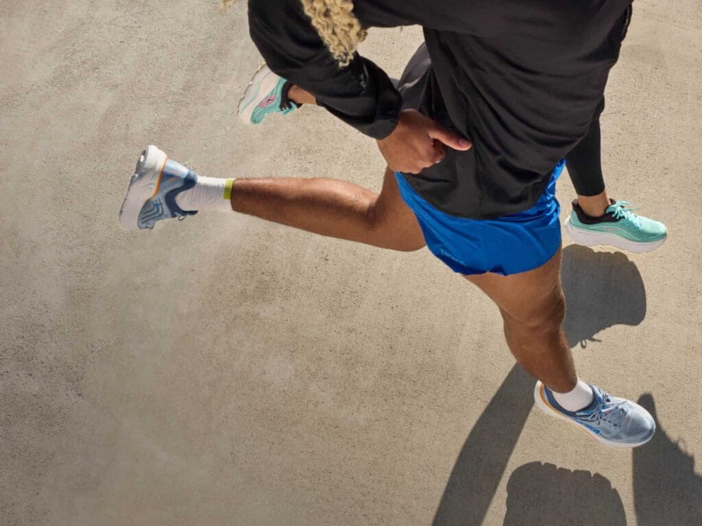 Vue de dessus de deux coureurs en pleine foulée sur le bitume, portant des chaussures de running Hoka (modèles Bondi et Clifton) et une tenue de sport