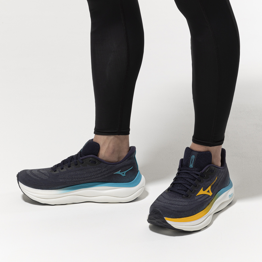 Coureur portant les Mizuno Wave Sky 9 bleu marine et jaune sur fond blanc, vue de profil et de face montrant le mesh et l'épaisseur de la semelle.