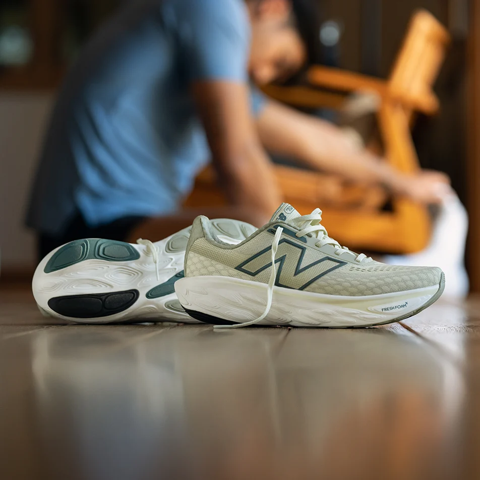 Paire de New Balance Fresh Foam X 1080 v14 beige posée sur un parquet, avec un coureur flou en arrière-plan qui se prépare