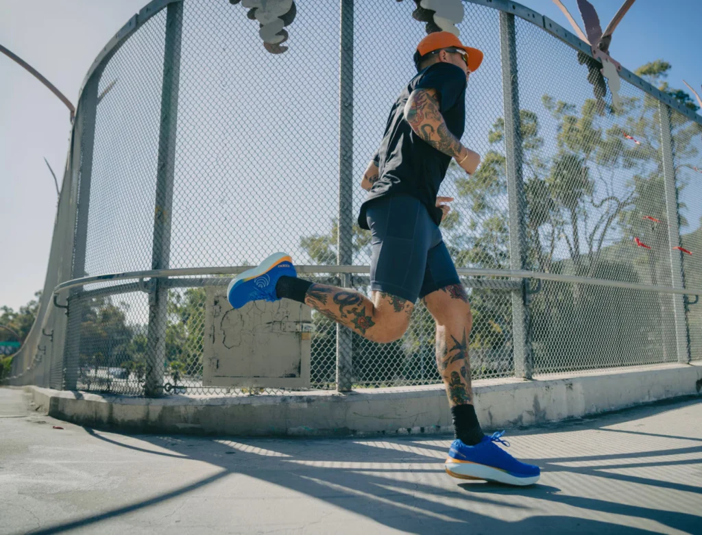 Coureur tatoué en pleine foulée sur un pont ensoleillé, portant une paire de Topo Athletic Atmos bleues à semelle épaisse blanche