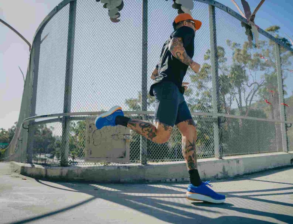 Coureur tatoué en pleine foulée sur un pont ensoleillé, portant une paire de Topo Athletic Atmos bleues à semelle épaisse blanche